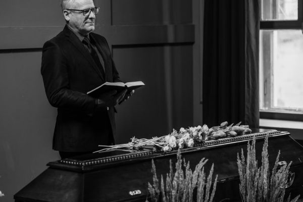 A solemn indoor funeral ceremony with a pastor standing beside a coffin.
